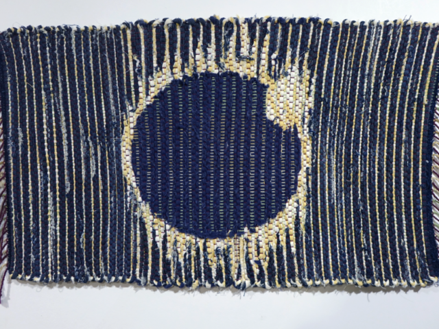 Woven rag wall hanging by fiber artist Petra Guenthner Johnson. Picture shows solar eclipse in navy denim with white and yellow outline with twisted fringe. Repurposed fabrics and cotton rug warp. 35x17"