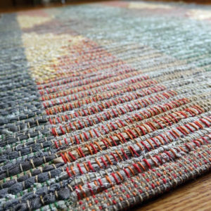 Detail of rag rug woven by fiber artist Petra Guenthner Johnson. Symmetrical geometric design in subtle grays, greens, yellows, and reds. Surface level detail of rolled edge and warp pattern. Made with repurposed materials and cotton rug warp. Aprroximate size 34x60".