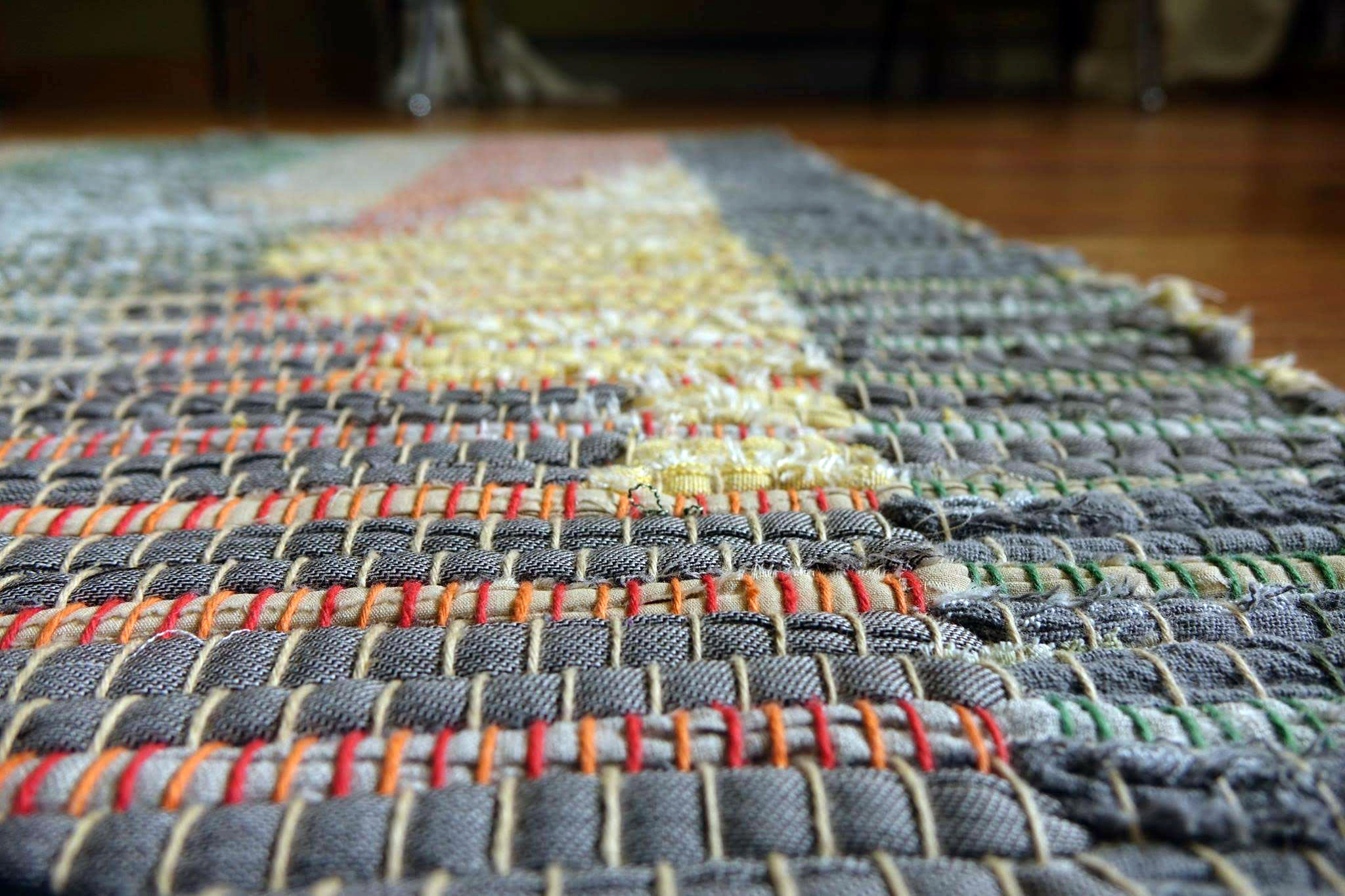 Detail of rag rug woven by fiber artist Petra Guenthner Johnson. Symmetrical geometric design in subtle grays, greens, yellows, and reds. Surface level detail showing fabric textures. Made with repurposed materials and cotton rug warp. Aprroximate size 34x60".