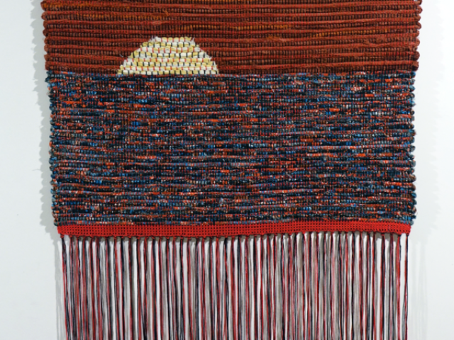 Woven fiber art by Petra Guenthner Johnson. Artwork depicts a yellow sun on a heavy red-orange sky setting on blue, red, orange, black mottled water. Long red and black fringe at the bottom.