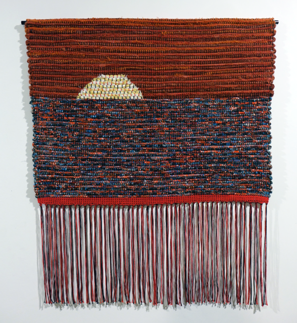 Woven fiber art by Petra Guenthner Johnson. Artwork depicts a yellow sun on a heavy red-orange sky setting on blue, red, orange, black mottled water. Long red and black fringe at the bottom.