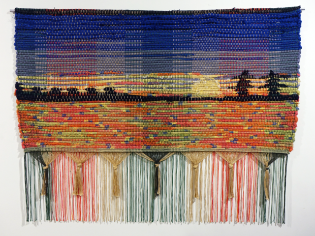 Woven rag wall hanging by fiber artist Petra Guenthner Johnson. Photo depicts shelterbelt silhouetted in black on a brightly colored sunset of red, yellow and blues. Repurposed fabrics and cotton rug warp. 34x22"