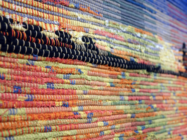 Detail of woven rag wall hanging by fiber artist Petra Guenthner Johnson. Photo depicts close-up of shelterbelt silhouetted in black on a brightly colored sunset of red, yellow and blues.