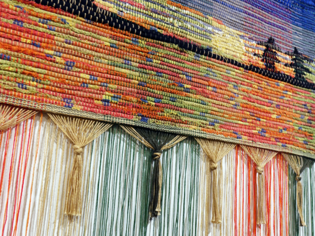 Detail of woven rag wall hanging by fiber artist Petra Guenthner Johnson. Photo depicts close-up of fringe with shelterbelt silhouetted in black on a brightly colored sunset of red, yellow and blues.