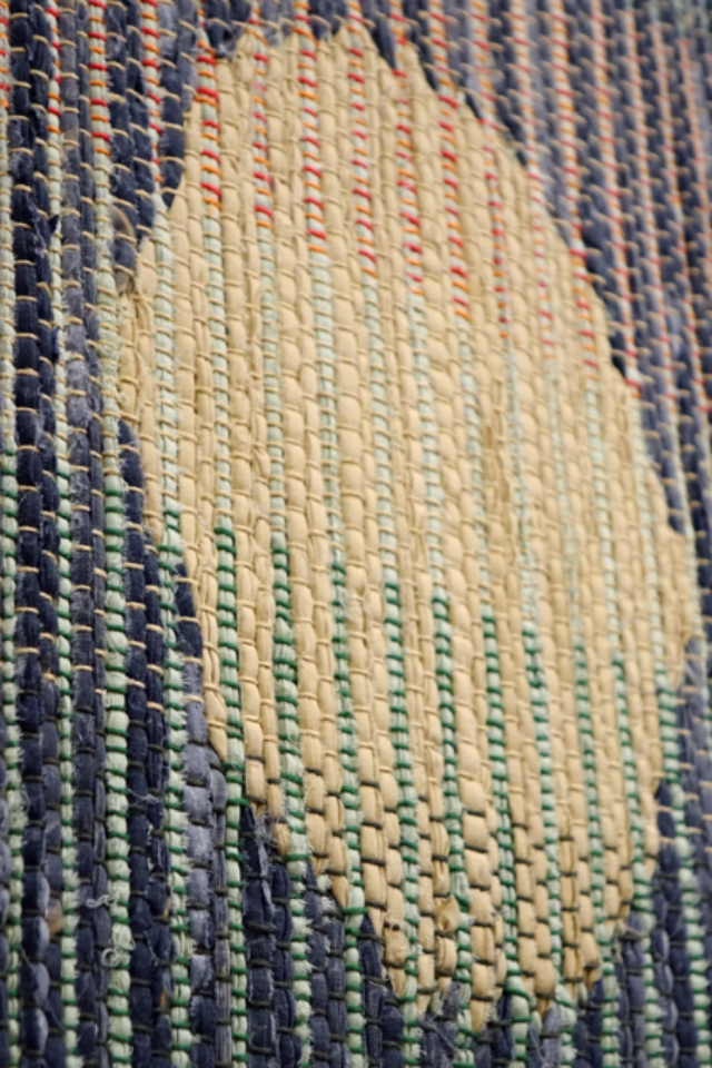 Woven rag wall hanging by fiber artist Petra Guenthner Johnson. Artwork is a close-up of a rag rug with neutral oval- shaped form up and right of center. It is on a gray background. Warp is in shades of green, orange and red. Heavy tassels drape down right side.