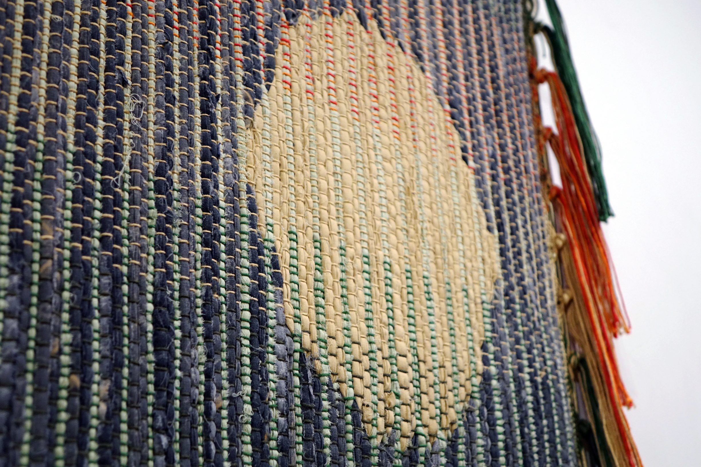 Woven rag wall hanging by fiber artist Petra Guenthner Johnson. Artwork is a close-up of a rag rug with neutral oval- shaped form up and right of center. It is on a gray background. Warp is in shades of green, orange and red. Heavy tassels drape down right side.