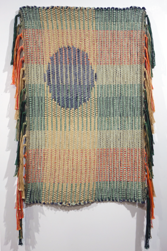 Woven rag wall hanging by fiber artist Petra Guenthner Johnson. Artwork is a rag rug with grey oval- shaped form up and left of center. It is on a neutral background. Warp is in shades of green, orange and red. Heavy tassels drape down right and left sides. Dimensions: 34x22"