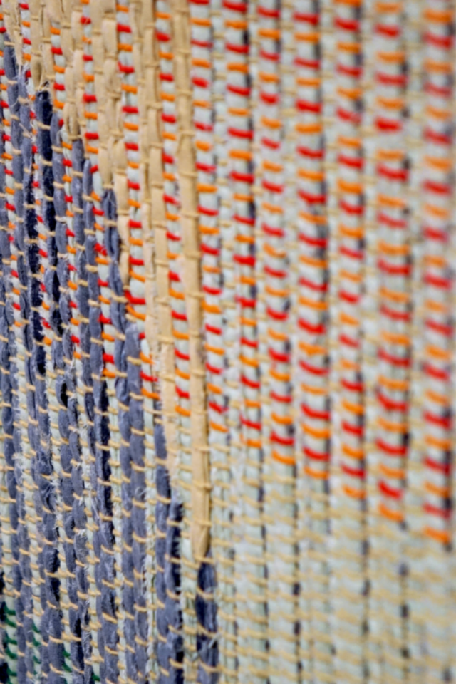 Detail of woven rag wall hanging by fiber artist Petra Guenthner Johnson. Close-up at an angle of a rag rug with grey oval- shaped form up and left of center. It is on a neutral background. Warp is in shades of green, orange and red. Heavy tassels drape down left side.