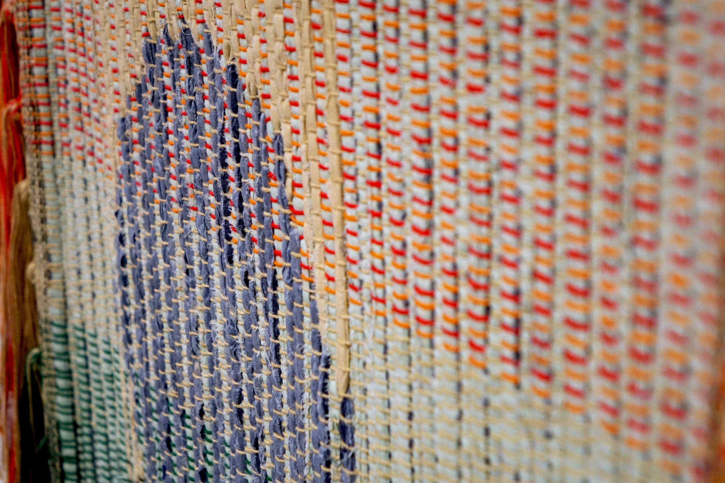 Detail of woven rag wall hanging by fiber artist Petra Guenthner Johnson. Close-up at an angle of a rag rug with grey oval- shaped form up and left of center. It is on a neutral background. Warp is in shades of green, orange and red. Heavy tassels drape down left side.