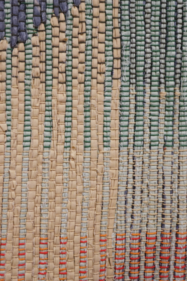 Detail of woven rag wall hanging by fiber artist Petra Guenthner Johnson. Close-up of a rag rug with grey form up and left of center. It is on a neutral background. Warp is in shades of green, orange and red.