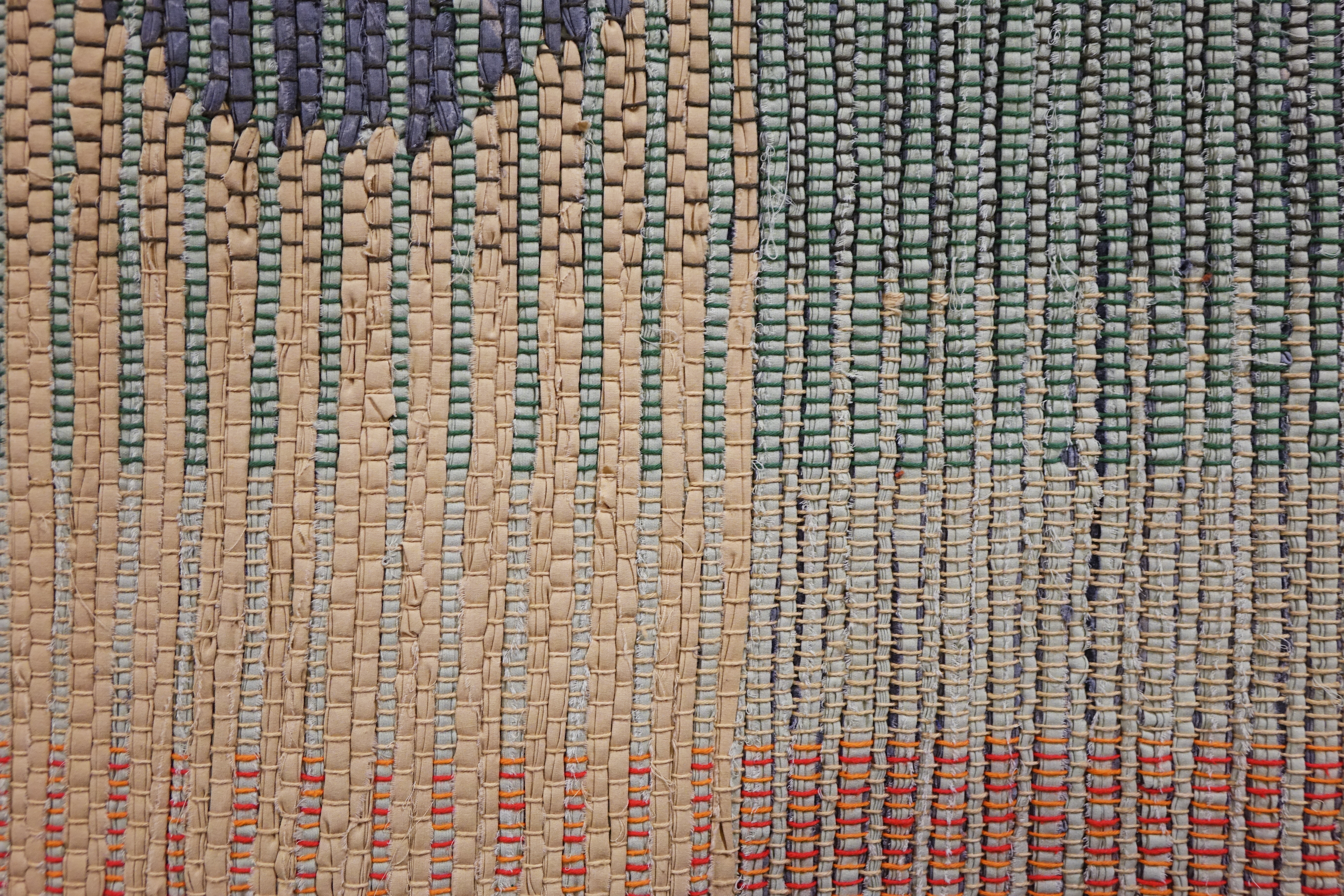 Detail of woven rag wall hanging by fiber artist Petra Guenthner Johnson. Close-up of a rag rug with grey form up and left of center. It is on a neutral background. Warp is in shades of green, orange and red.