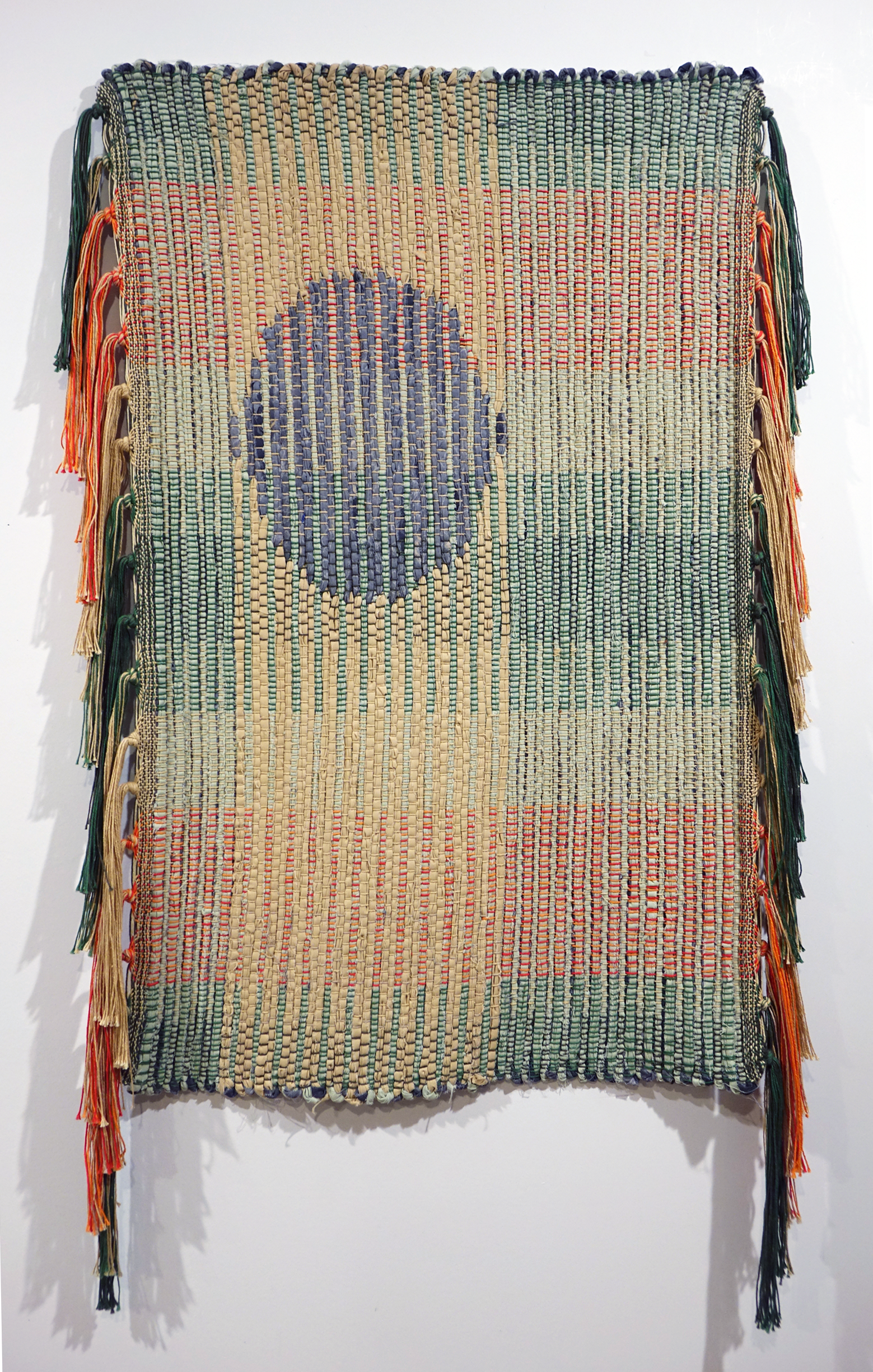 Woven rag wall hanging by fiber artist Petra Guenthner Johnson. Artwork is a rag rug with grey oval- shaped form up and left of center. It is on a neutral background. Warp is in shades of green, orange and red. Heavy tassels drape down right and left sides. Dimensions: 34x22"