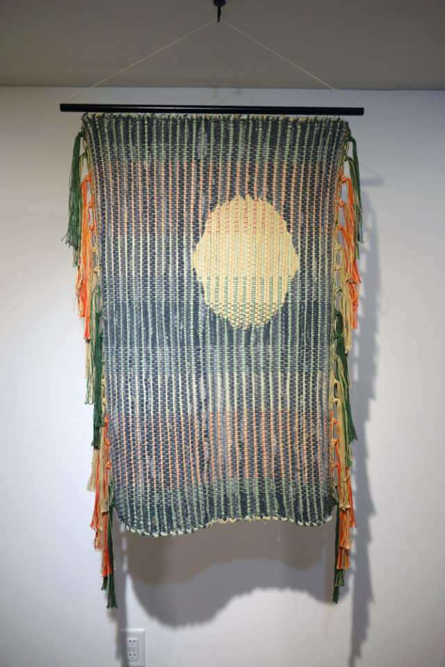 Woven rag wall hanging by fiber artist Petra Guenthner Johnson. Artwork is a rag rug with off-white oval- shaped form up and left of center. It is on a gray background. Warp is in shades of green, orange and red. Heavy tassels drape down right and left sides. Dimensions: 34x22"