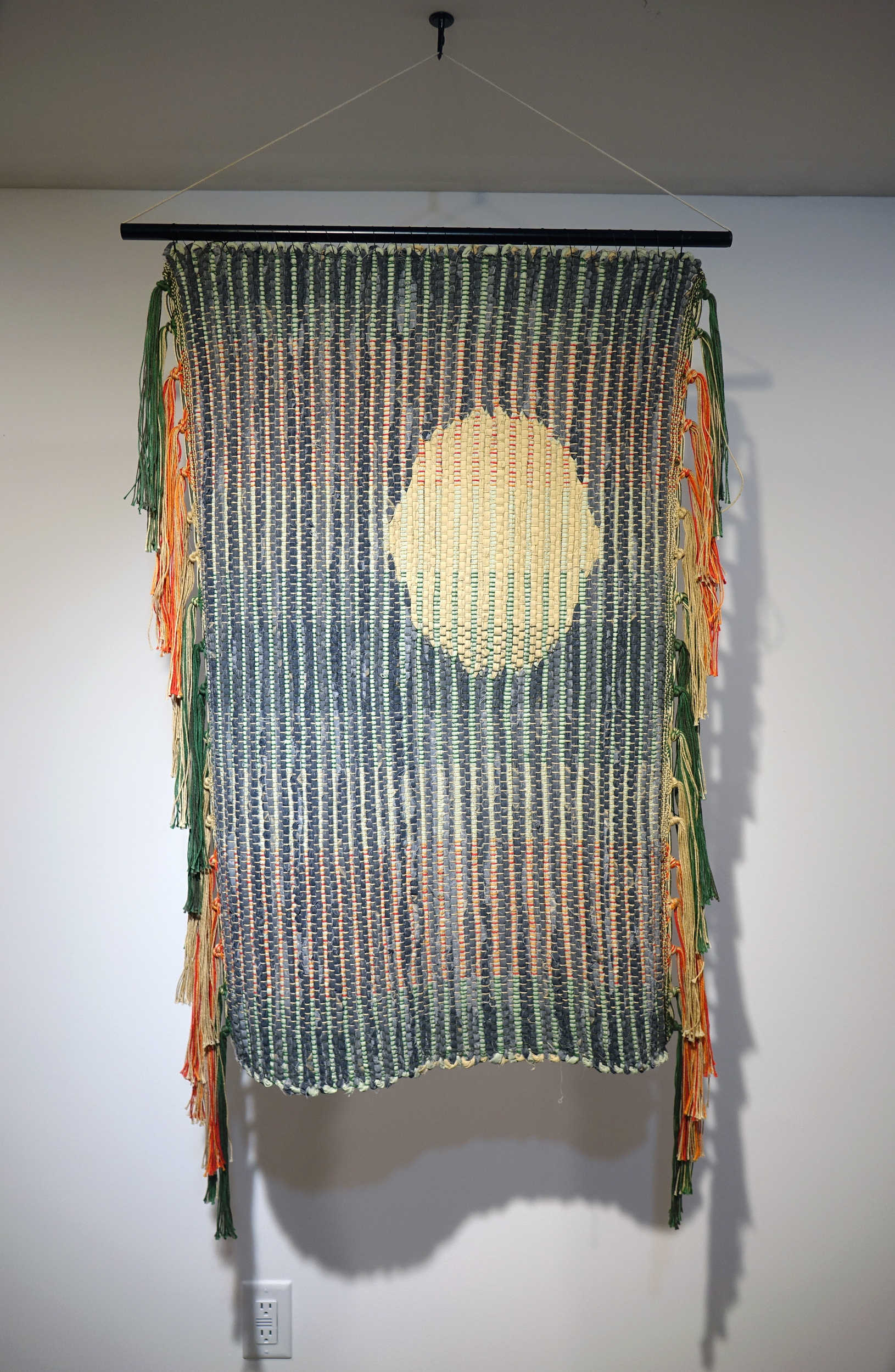 Woven rag wall hanging by fiber artist Petra Guenthner Johnson. Artwork is a rag rug with off-white oval- shaped form up and left of center. It is on a gray background. Warp is in shades of green, orange and red. Heavy tassels drape down right and left sides. Dimensions: 34x22"