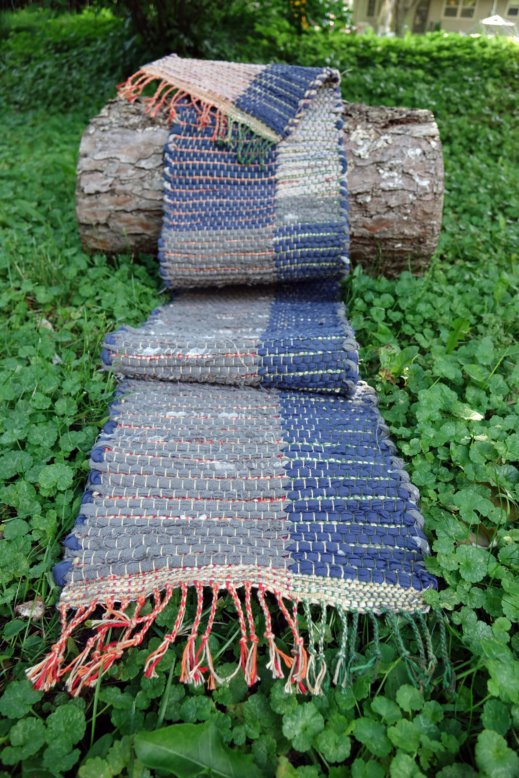 Rag rug woven by fiber artist Petra Guenthner Johnson. Table runner draped over small log, design is in four quadrants; alternating dark blue and gray/taupe with red and green details.