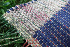 Detail of rag rug woven by fiber artist Petra Guenthner Johnson. Rug on green grass woven one side of rug is dark blue the other side is taupe. This detail show a close-up of fabric texture and twisted fringes. Made with repurposed materials and cotton rug warp. Approximate size 10x52".