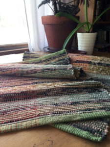 Rag rugs bunched on table. Shows light green woven, rolled hems. Rugs are multi-colored in yellows, blacks, red, orange and green.