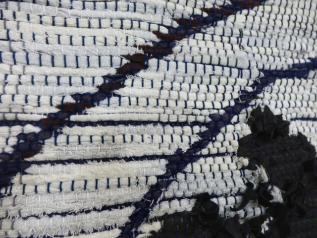 Close-up shot of woven rag wall hanging by fiber artist Petra Guenthner Johnson. Artwork has white background with black form in lower right corner. Navy and purple lines travel from bottom left of image to upper right. 34x22"